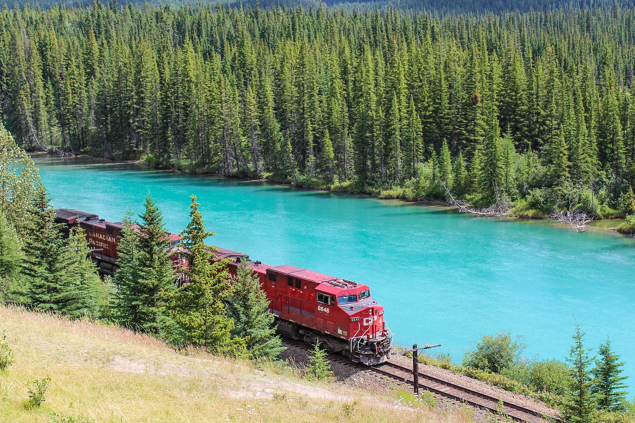 A red Canadian Pacific train travels along a track next to a bright turquoise river surrounded by pine forests.