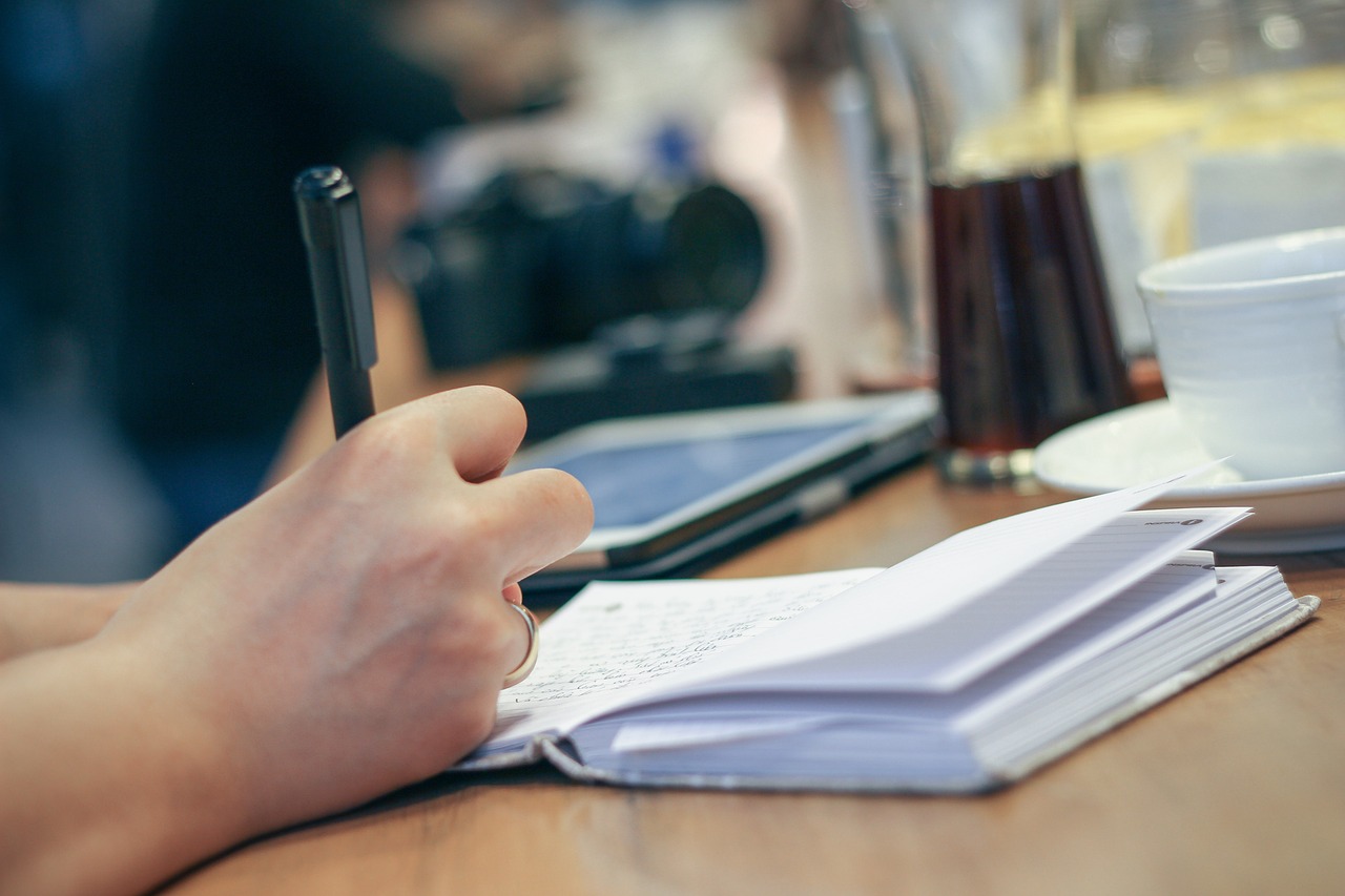 Close-up of a hand writing in an open notebook with a pen on a wooden table.