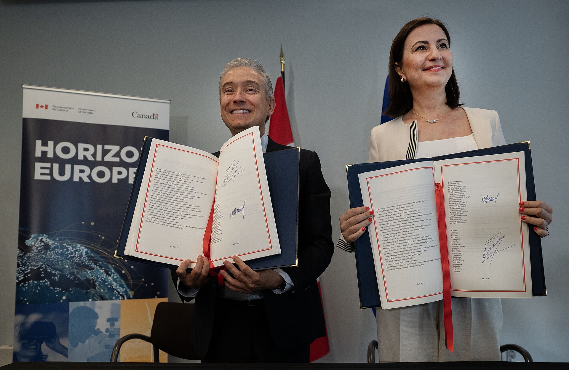 A man and a woman hold open large documents with red ribbons, standing in front of a &ldquo;Government of Canada&rdquo; banner.