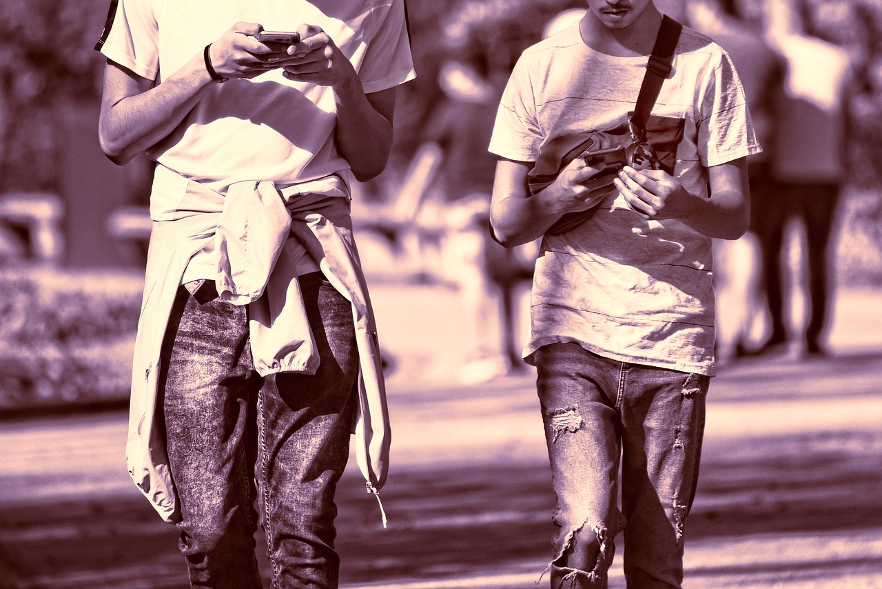 Two young men walk outdoors, looking at their smartphones, in a purple-toned image.