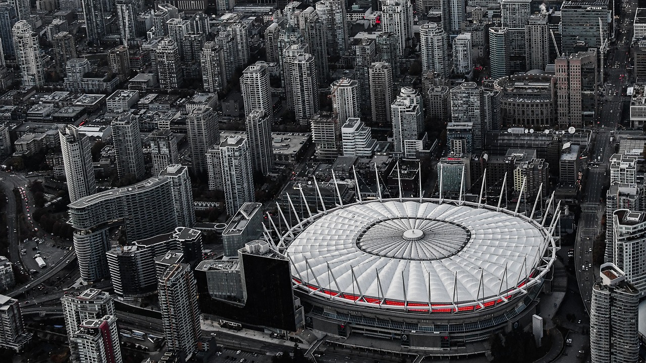 Aerial view of a city with many tall buildings surrounding a large, round stadium with a white roof.