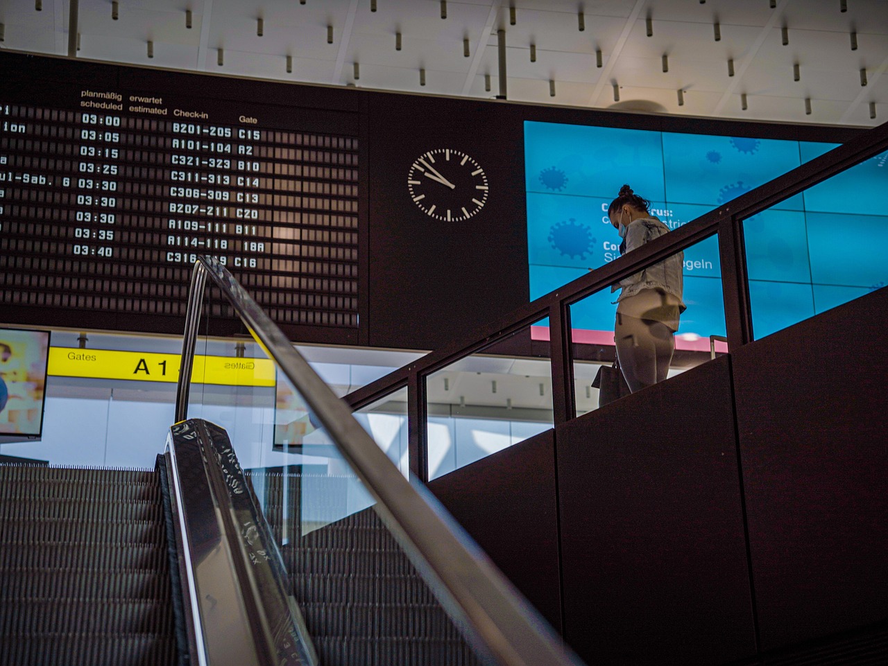 An escalator in an airport terminal with a large digital departure board and another screen displaying "Coronavirus" information.