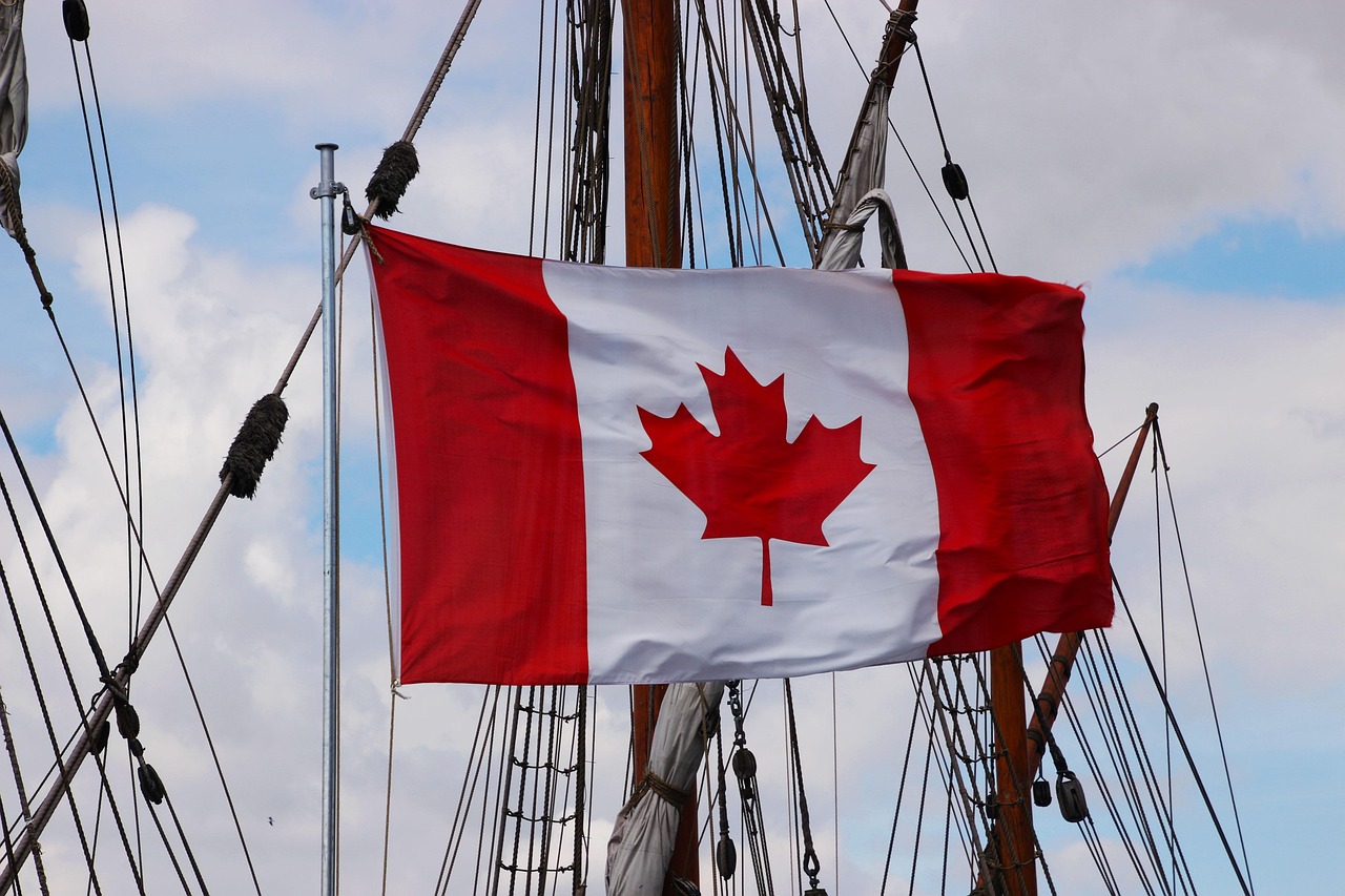 Canadian flag with a red maple leaf flying from a flagpole, with ship rigging and masts in the background.