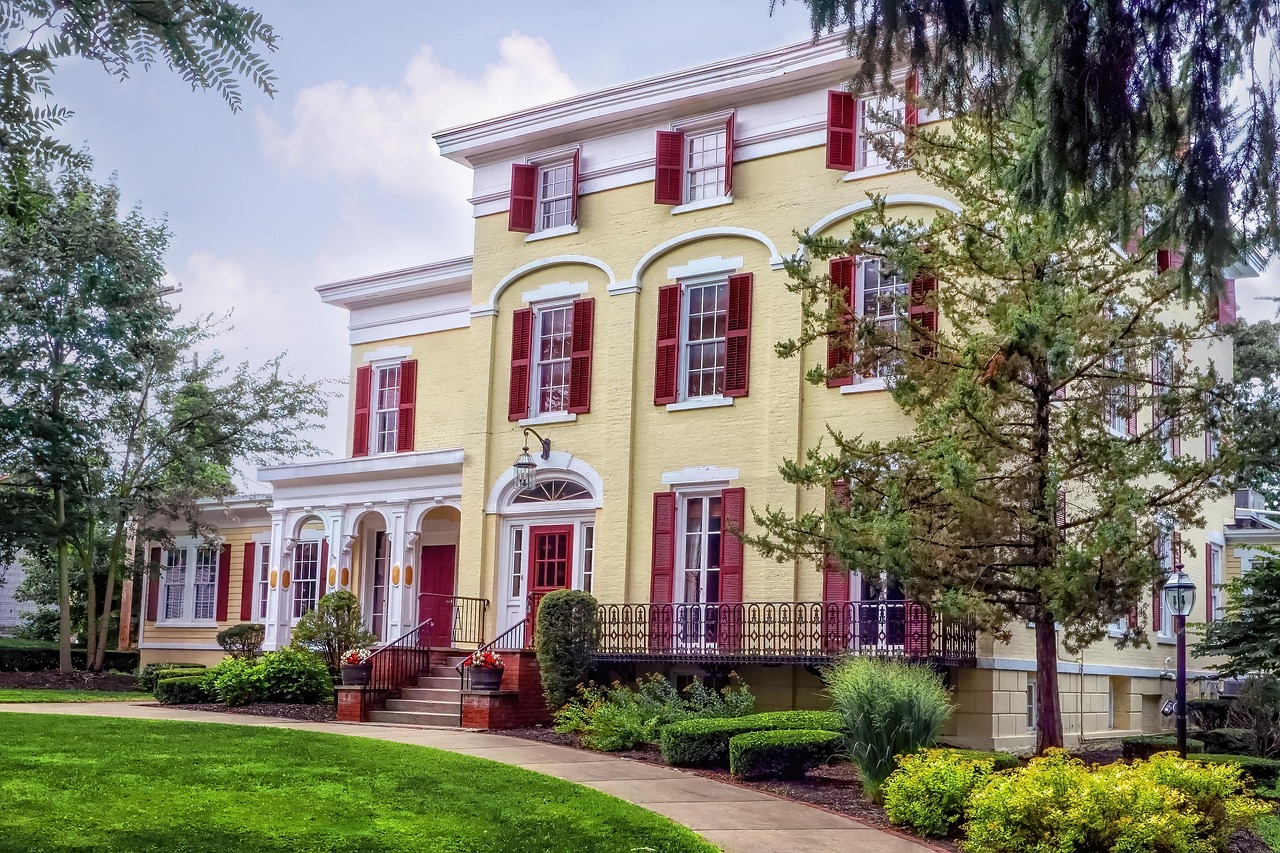 A large, two-story yellow house with red shutters, white trim, a green lawn, and trees.