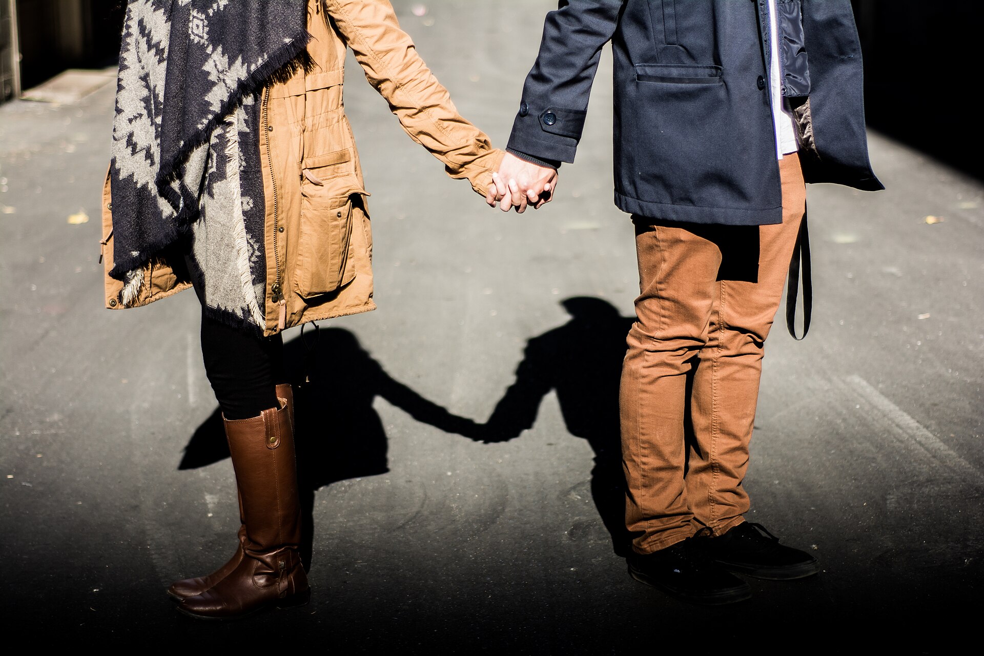 Two people holding hands, seen from the waist down, standing on a dark paved surface.