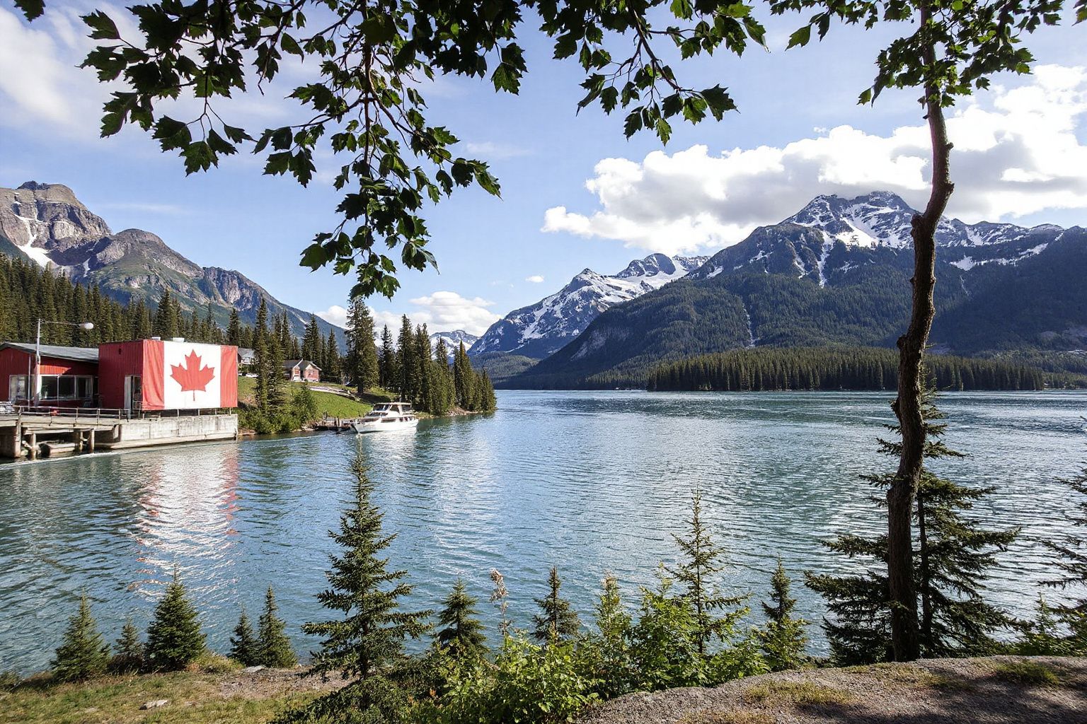 Scenic view of a lake with a red building displaying a large Canadian flag, surrounded by evergreen trees and snow-capped mountains.