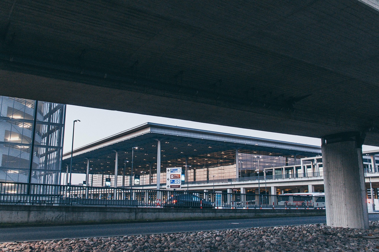 Modern airport terminal building with glass facade, seen from under a concrete overpass, with cars and parking signs.