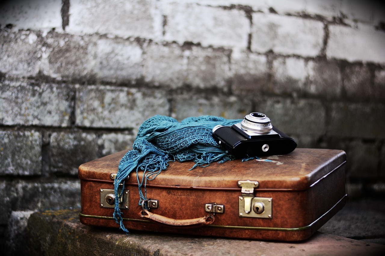 A brown vintage suitcase with a teal scarf and an old film camera on top, against a blurred brick wall.