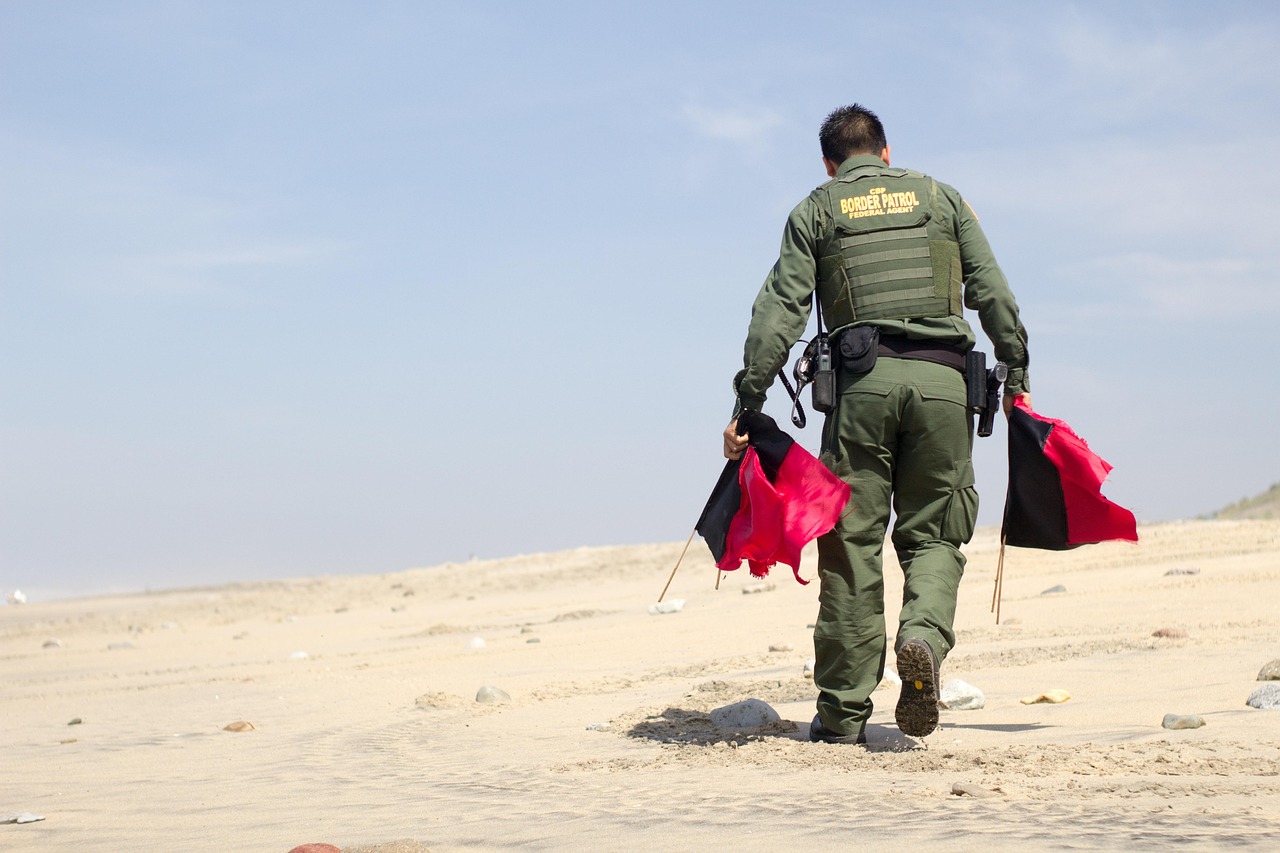 A person in a &ldquo;BORDER PATROL&rdquo; uniform walks on a sandy beach holding red and black flags.
