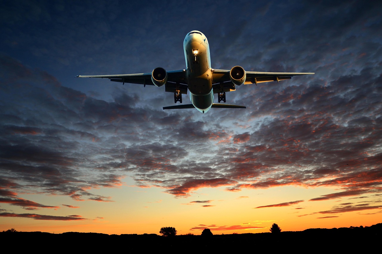 Large passenger airplane with landing gear down flying against a dramatic sunset sky over a dark landscape.