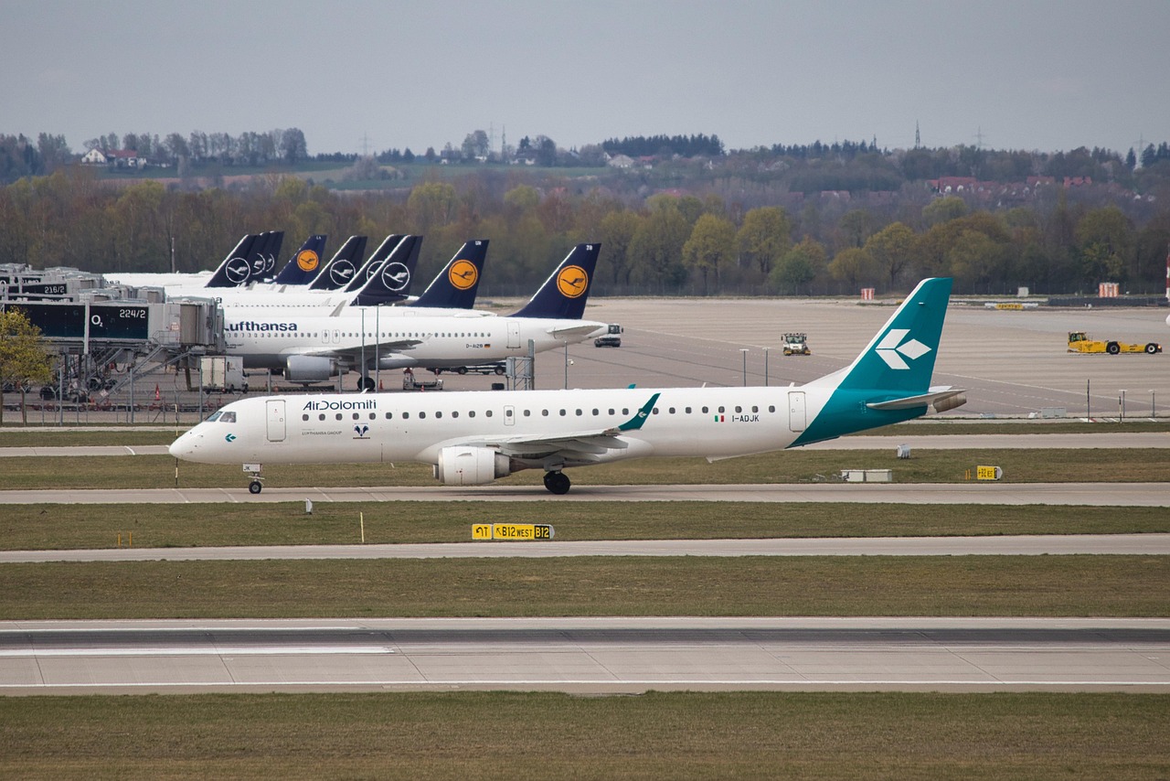 An Air Dolomiti aircraft taxis on an airport tarmac with several Lufthansa planes parked at gates in the background.