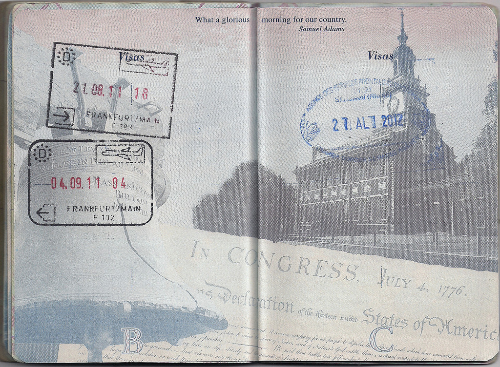 Open passport pages with various entry stamps, including Frankfurt and a blue circular stamp over a faint image of Independence Hall.