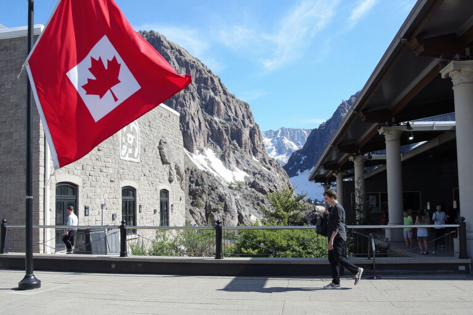 A Canadian flag flies in front of a stone building and snow-capped mountains, with people walking on a paved path.