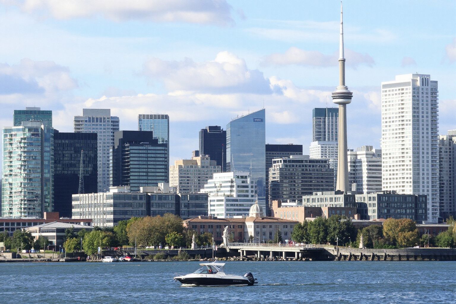 City skyline with a tall tower and modern buildings, a boat on the water, under a partly cloudy sky.