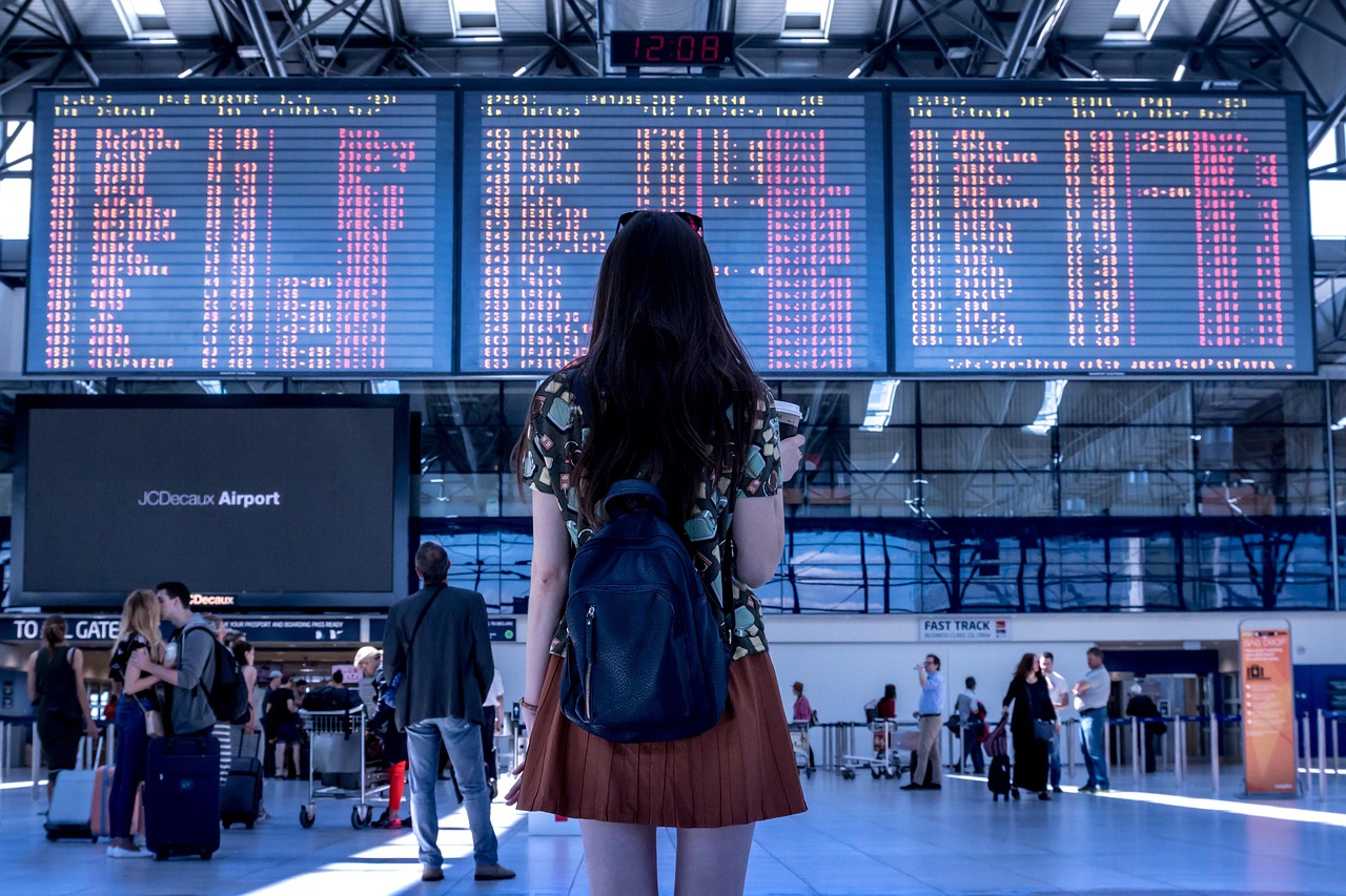 A person with a backpack stands in front of a large airport departure board displaying flight information.