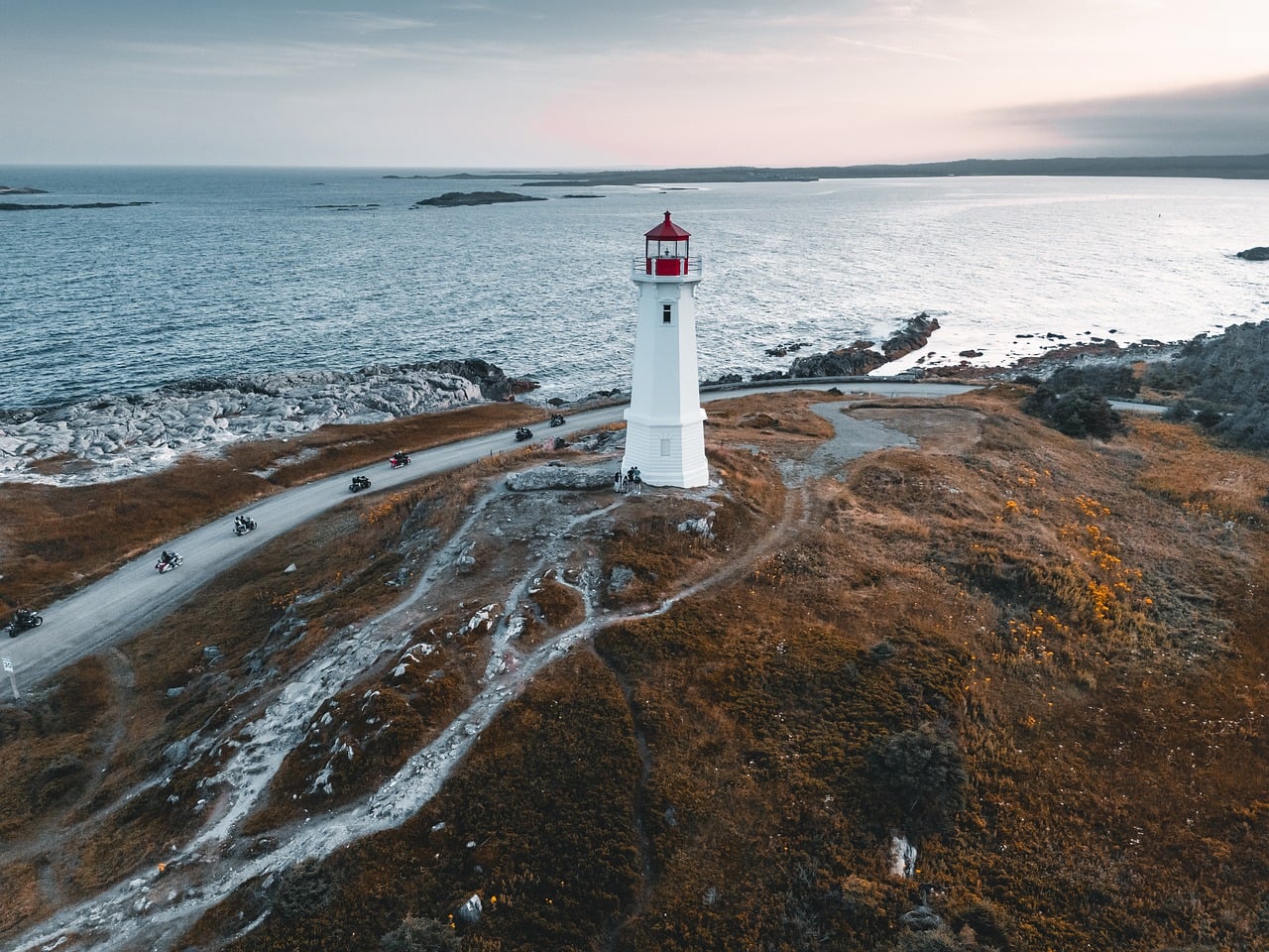 Aerial view of a white lighthouse with a red top on a rocky coast with motorcycles on a dirt road.