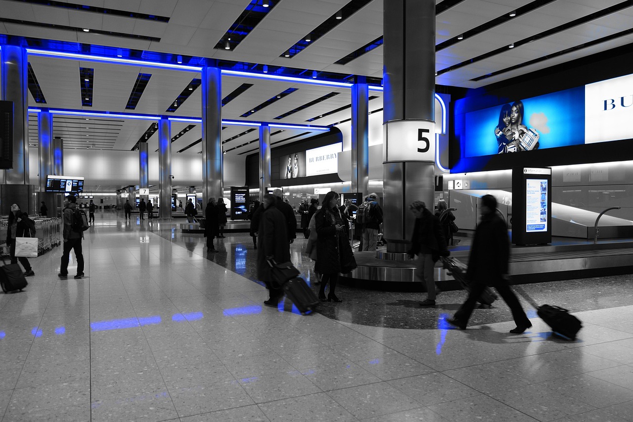 People walking with luggage in a modern airport terminal with blue lighting accents and a baggage claim carousel.
