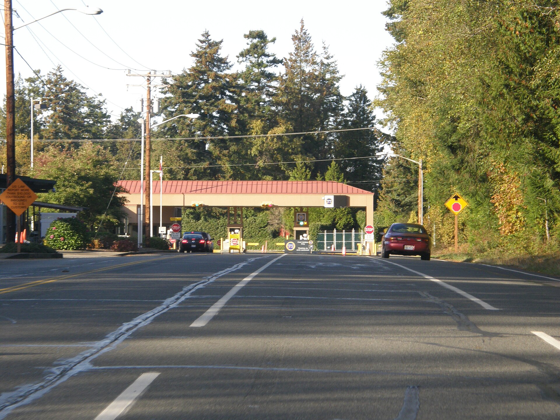 A two-lane road leading towards a building resembling a border crossing, flanked by evergreen trees.