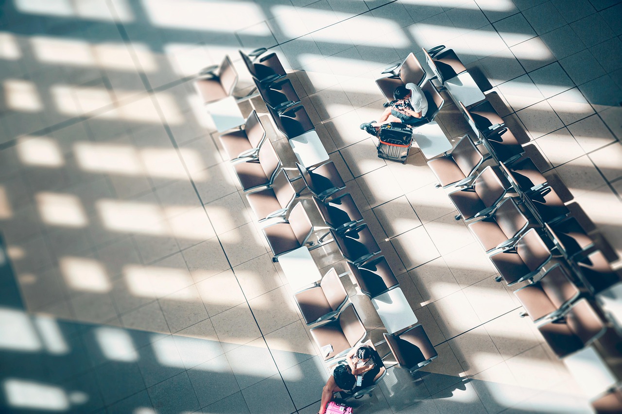 Overhead view of an airport waiting area with rows of chairs, two travelers with luggage, and strong window shadows.