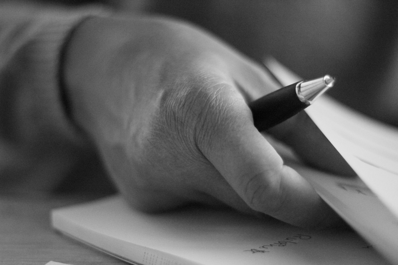 Close-up of a hand holding a pen over a notebook, in black and white.