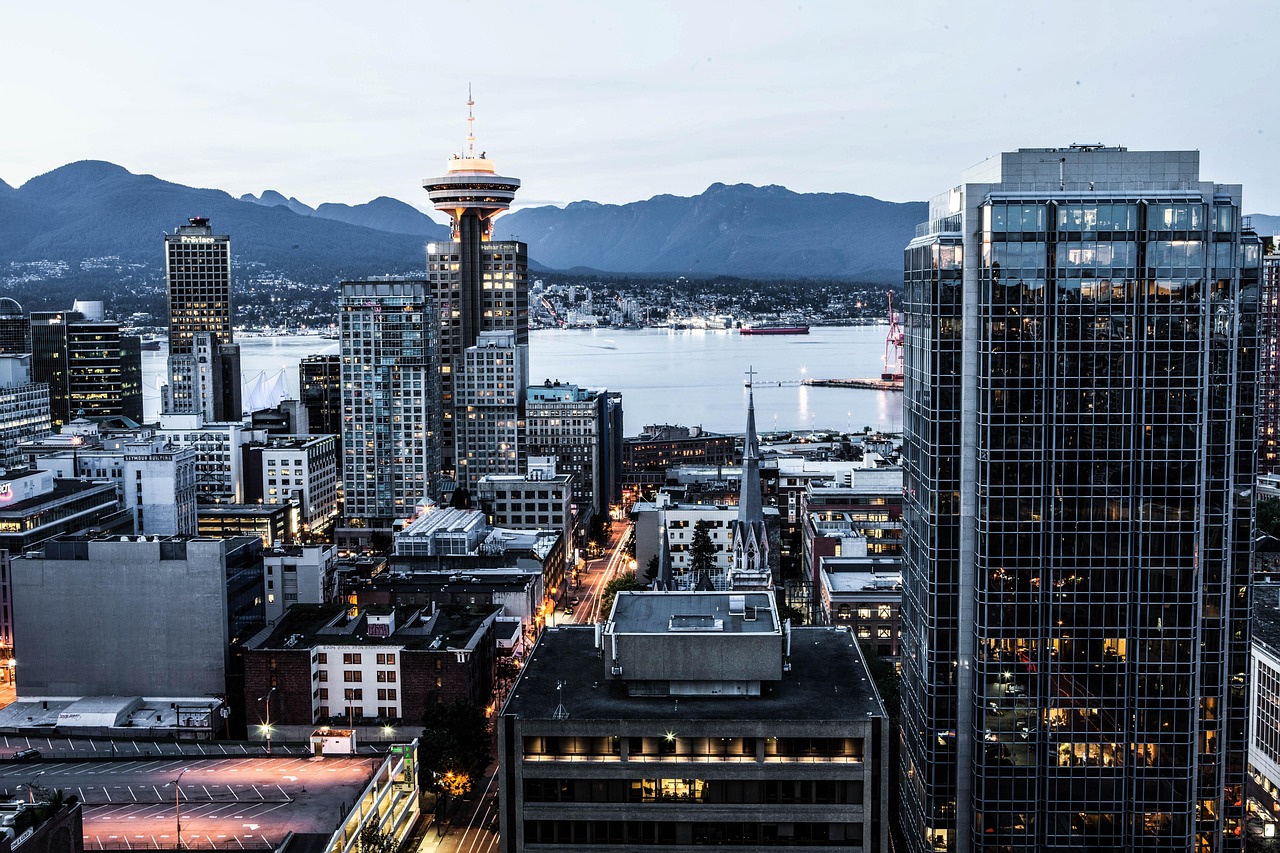 Cityscape at dusk with tall buildings, a prominent observation tower, a body of water, and mountains in the background.