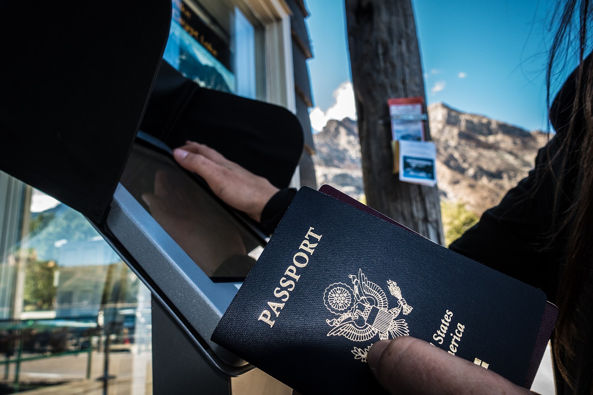 A hand holds a US passport near a self-service kiosk, with another hand touching the screen. Mountains in background.