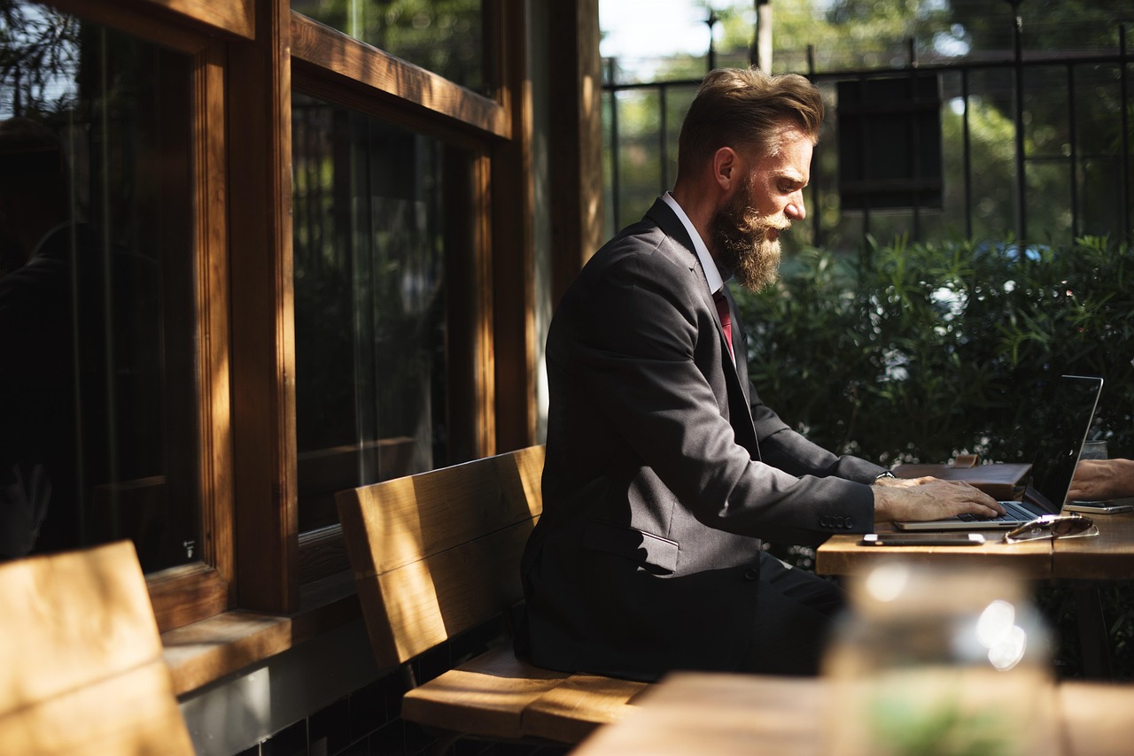 Bearded man in a suit typing on a laptop at an outdoor wooden table with sunglasses nearby.