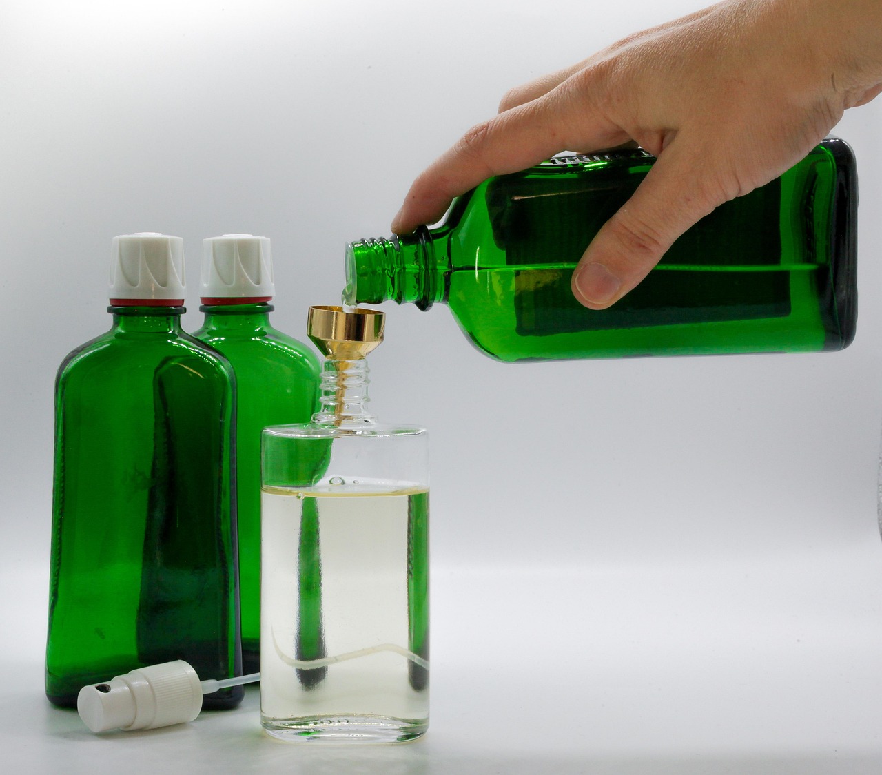 Hand pouring liquid from a green bottle through a funnel into a clear glass, with other bottles nearby.