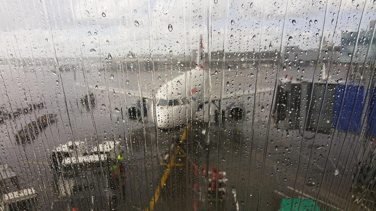 A commercial airplane at an airport gate seen through a rain-streaked window with ground equipment on the tarmac.