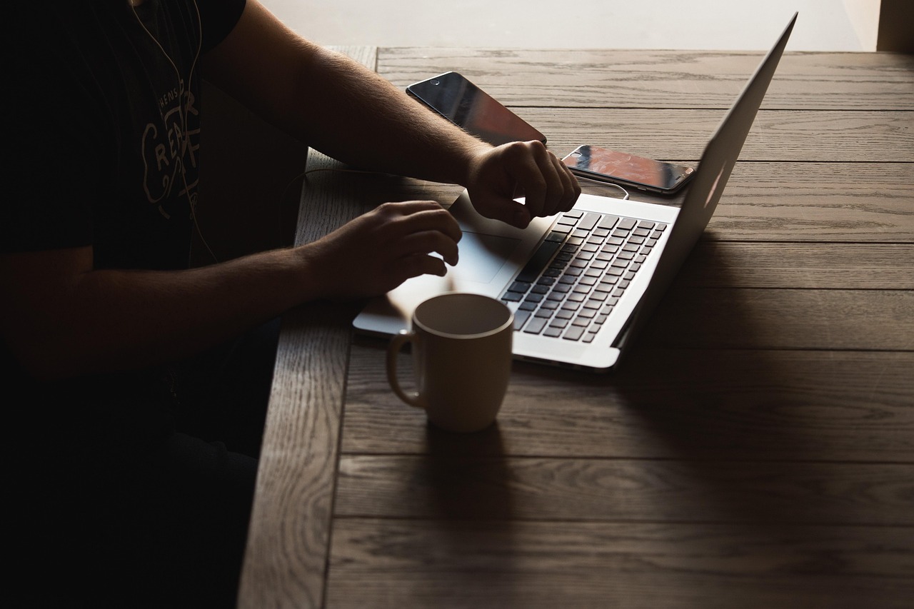 Person typing on a silver laptop at a wooden table with a mug and two smartphones.