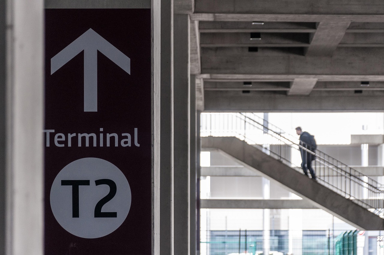A dark red sign with a white arrow and "Terminal T2" text, with a person walking up concrete stairs in the background.