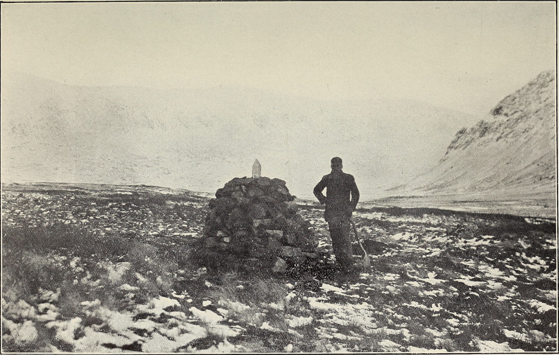 Black and white photo of a person standing by a stone cairn on a snowy, rocky mountain landscape.