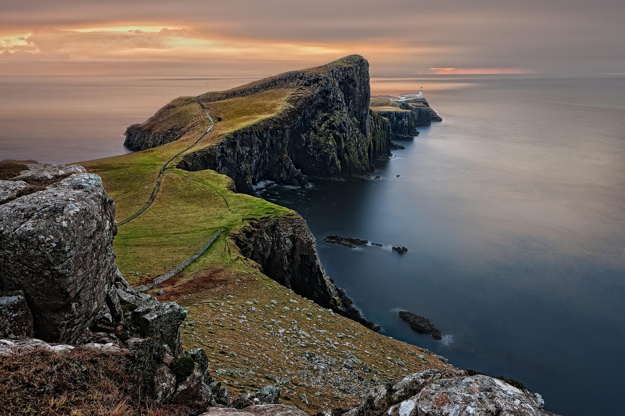 Dramatic coastal landscape with a winding path along cliffs leading to a white lighthouse at sunset.