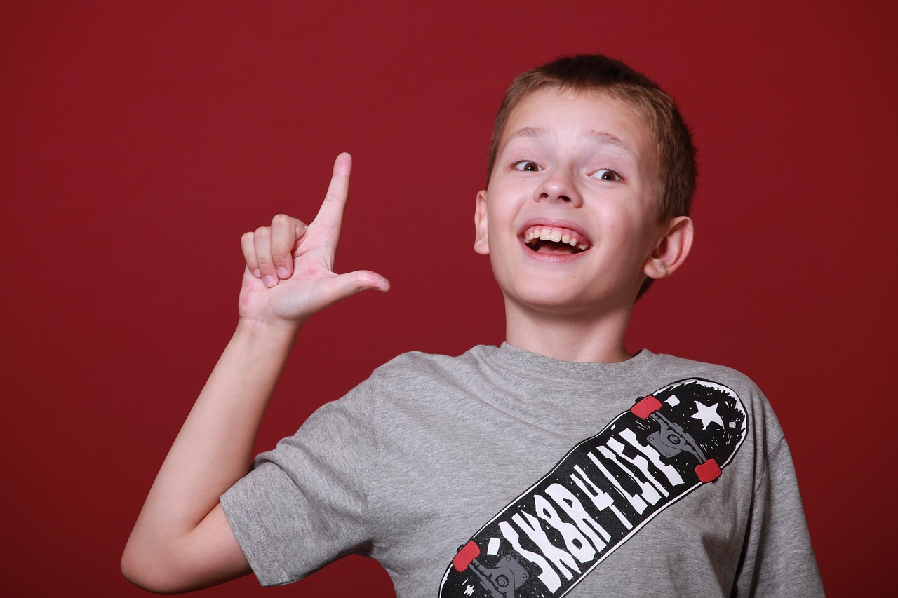 A smiling boy in a grey t-shirt with a skateboard graphic makes an &ldquo;L&rdquo; shape with his hand against a red background.
