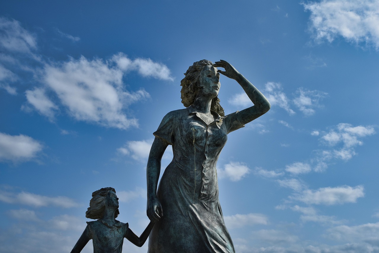 A low-angle bronze statue of a woman shading her eyes and a smaller figure against a blue sky with clouds.