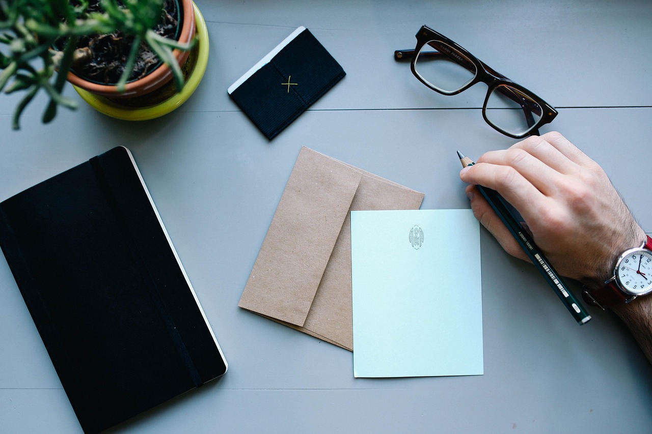 Hand holding a pencil over a light blue note card, next to a brown envelope, black notebook, and glasses.