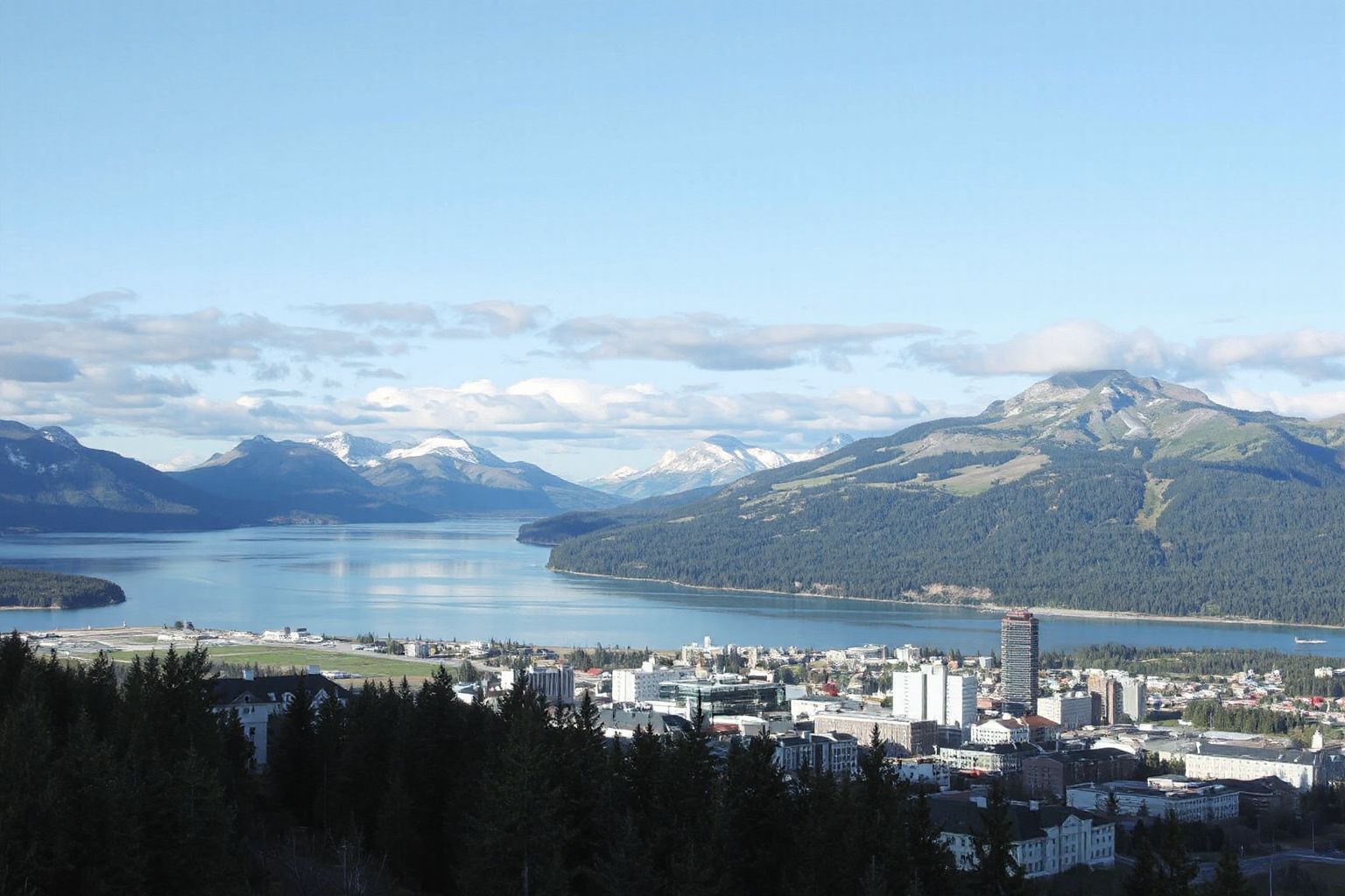 Panoramic view of a city by a large body of water with an airport, surrounded by forested mountains and a blue sky.