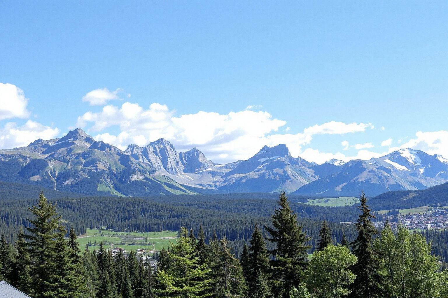 Panoramic view of a mountain range with snow-capped peaks, dense evergreen forests, a green valley, and a small town under a blue sky.