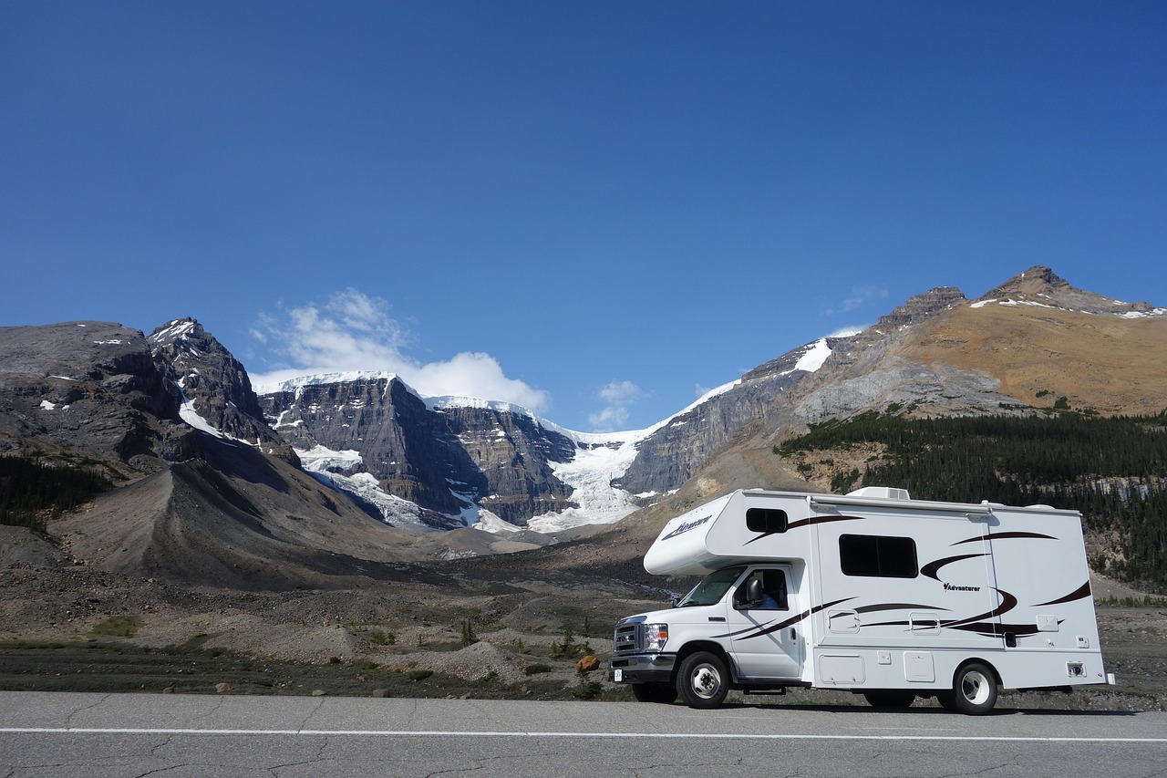 White RV parked on a paved road with snow-capped mountains and glaciers under a clear blue sky.