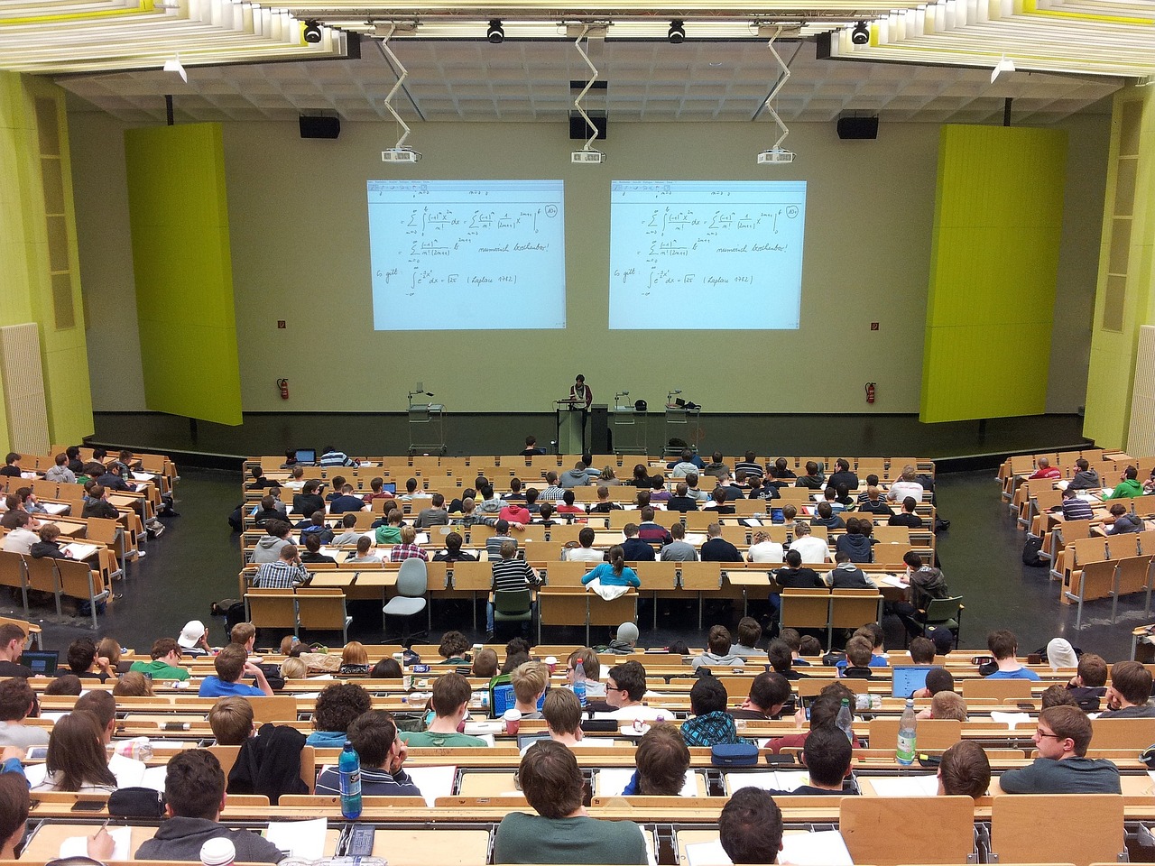 Overhead view of a large lecture hall filled with students, with a lecturer and two screens displaying equations.