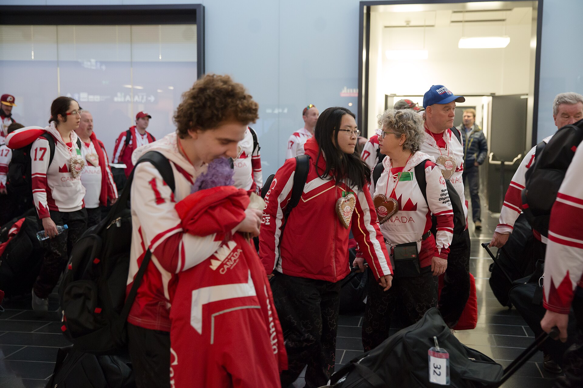 Group of people in red and white &ldquo;Canada&rdquo; jackets with luggage walking through an airport terminal.