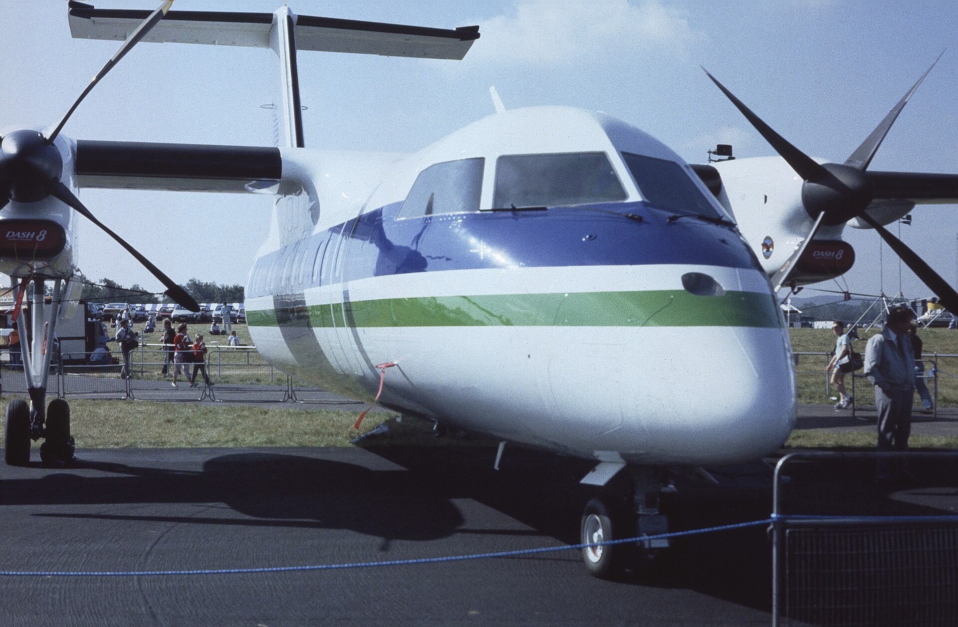 Front-side view of a white, blue, and green propeller plane (Dash 8) parked on an airfield with people in the background.