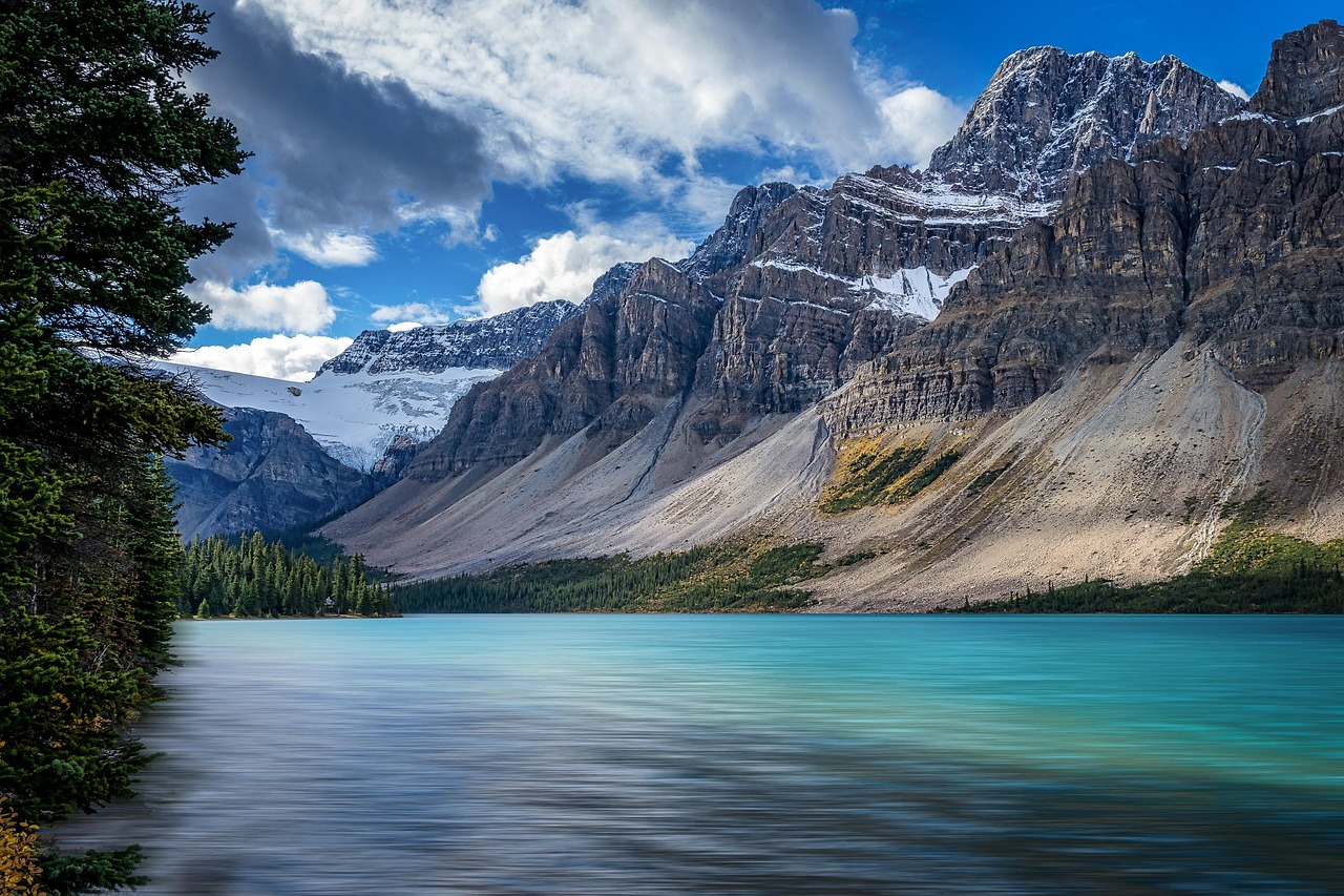 Turquoise lake with snow-capped mountains and evergreen forests under a dramatic cloudy sky.
