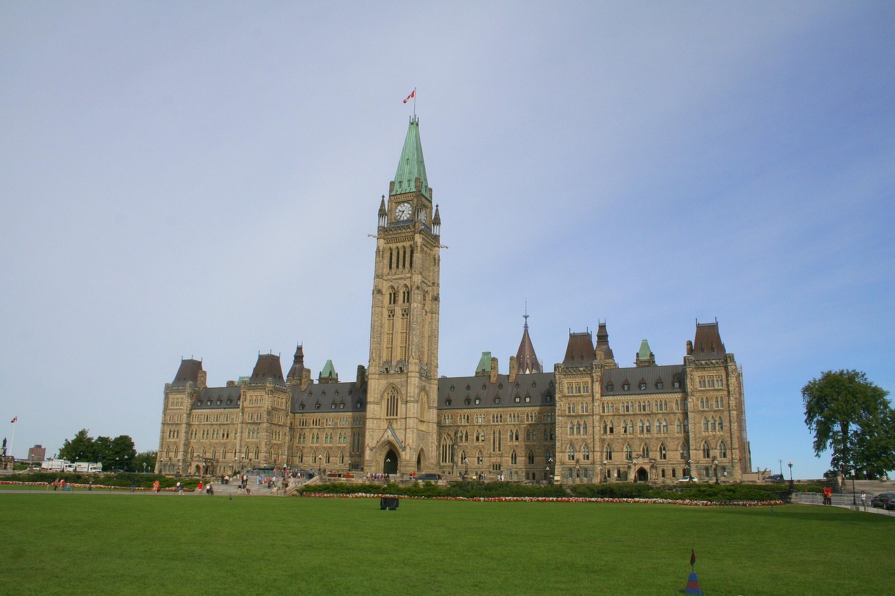 Wide shot of a large, ornate stone building with a tall clock tower and a Canadian flag, fronted by a green lawn.