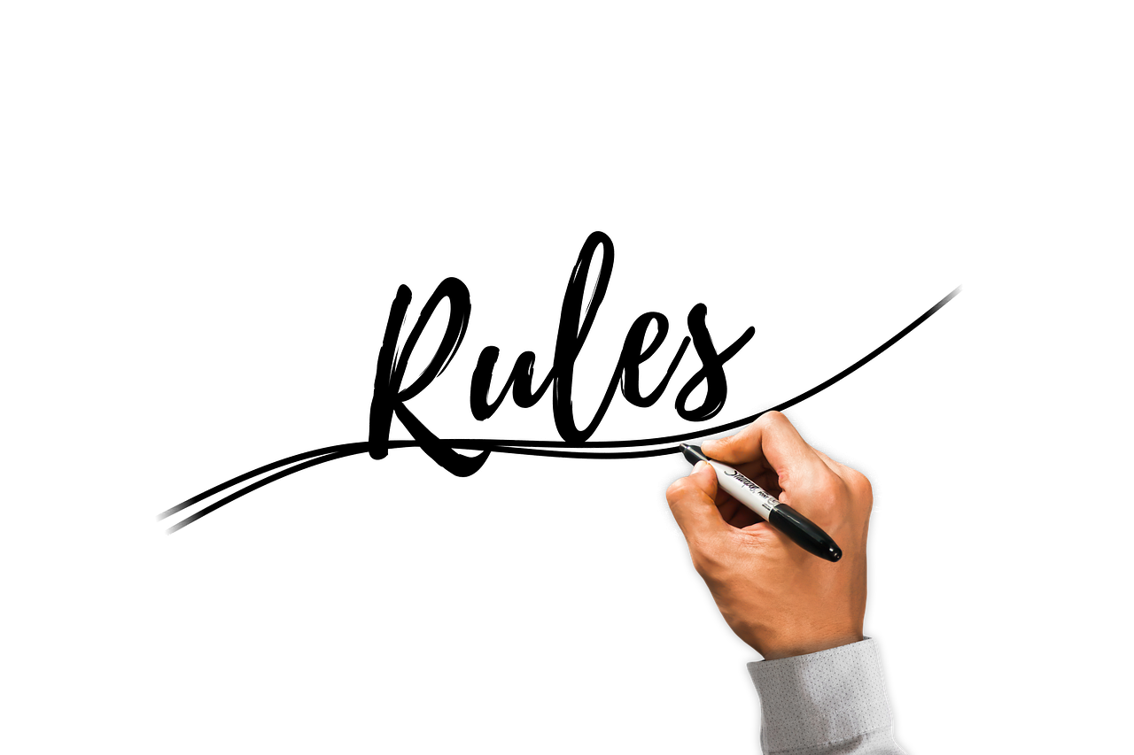 A hand holding a black marker pen drawing a line under the word “Rules” written in cursive on a white background.