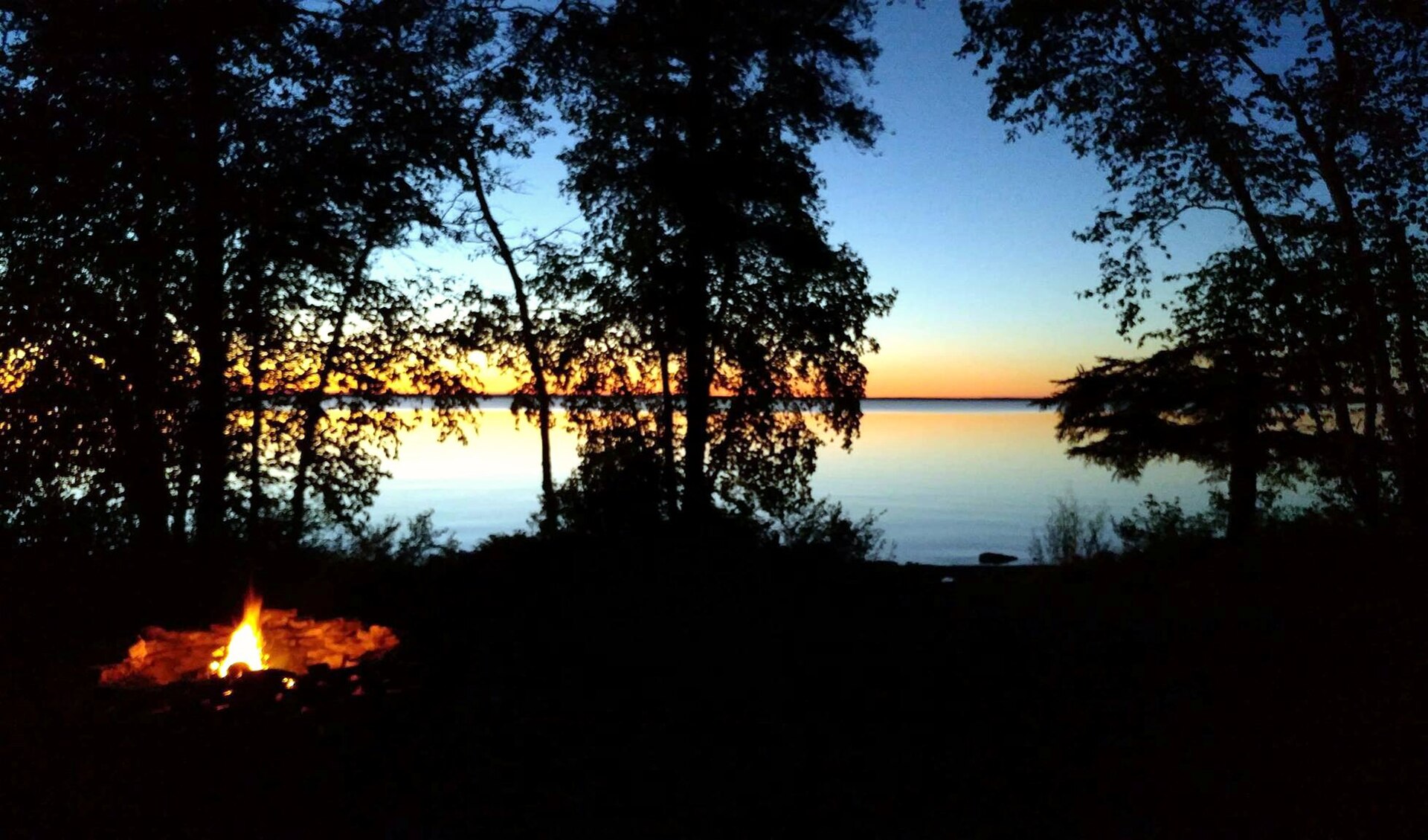 Silhouetted trees frame a lake at sunset or sunrise with a small campfire in the foreground.