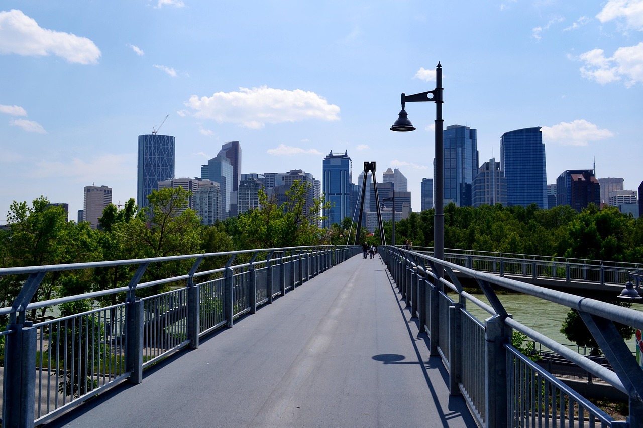A long pedestrian bridge leads towards a city skyline with modern high-rise buildings and green trees under a blue sky.