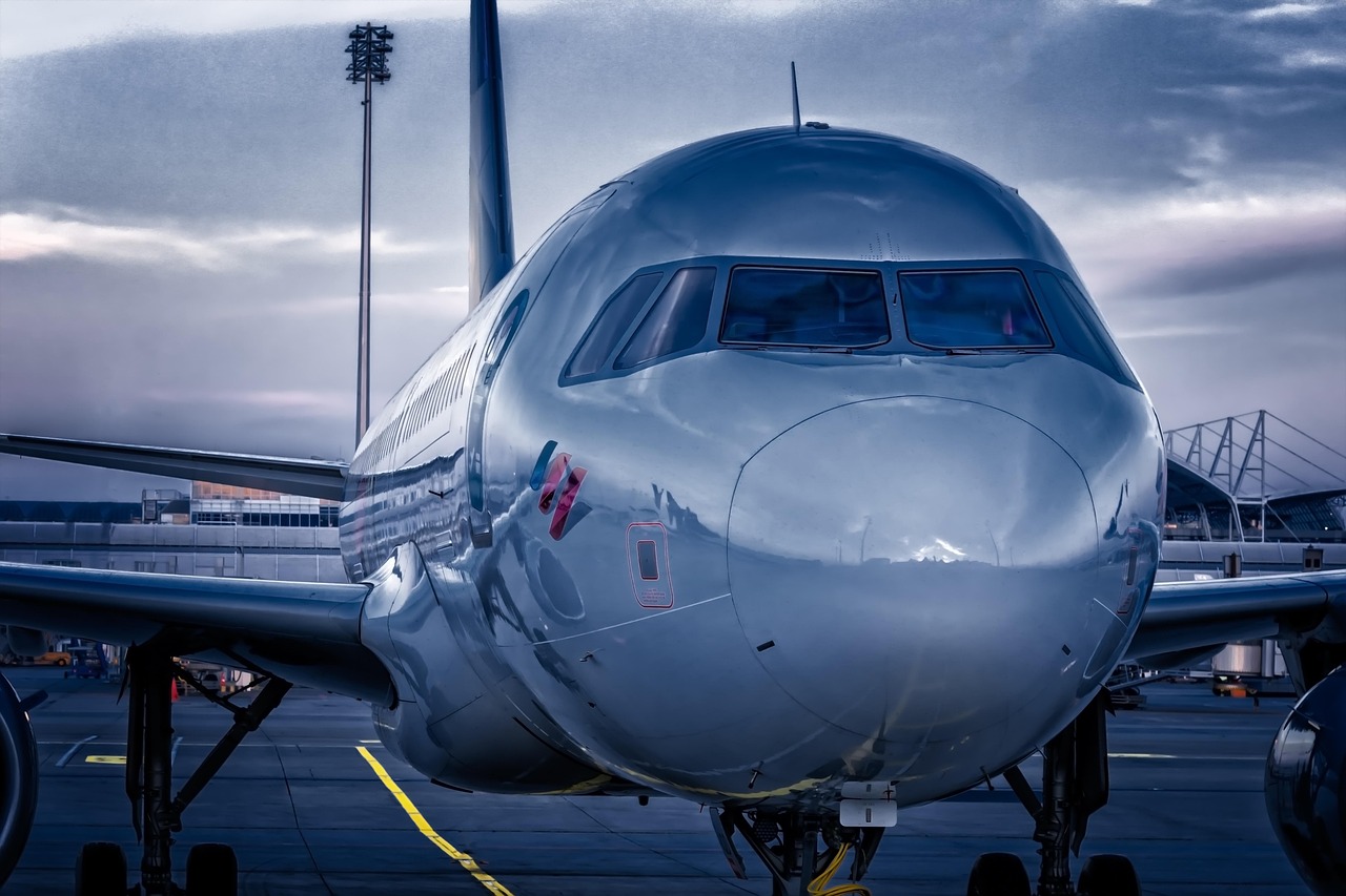 Front view of a metallic grey passenger airplane parked on an airport tarmac under a cloudy sky.