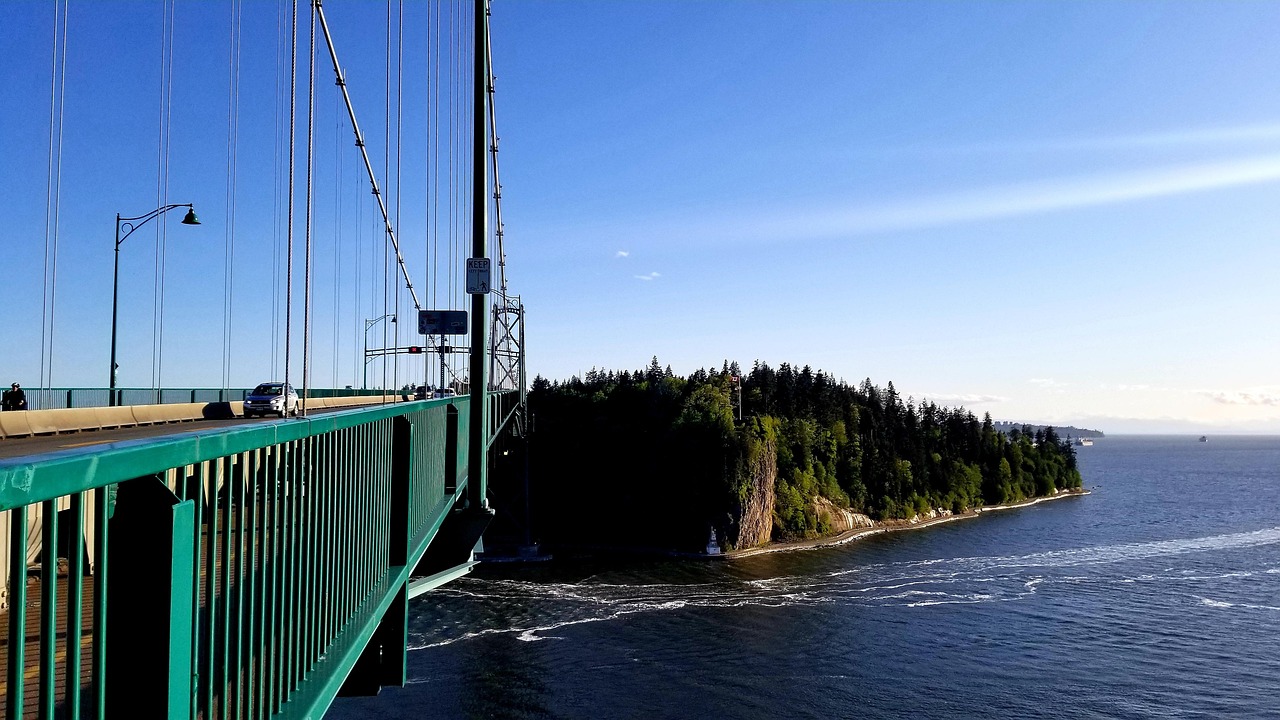 A large suspension bridge with green railings over water next to a forested cliff under a clear blue sky.