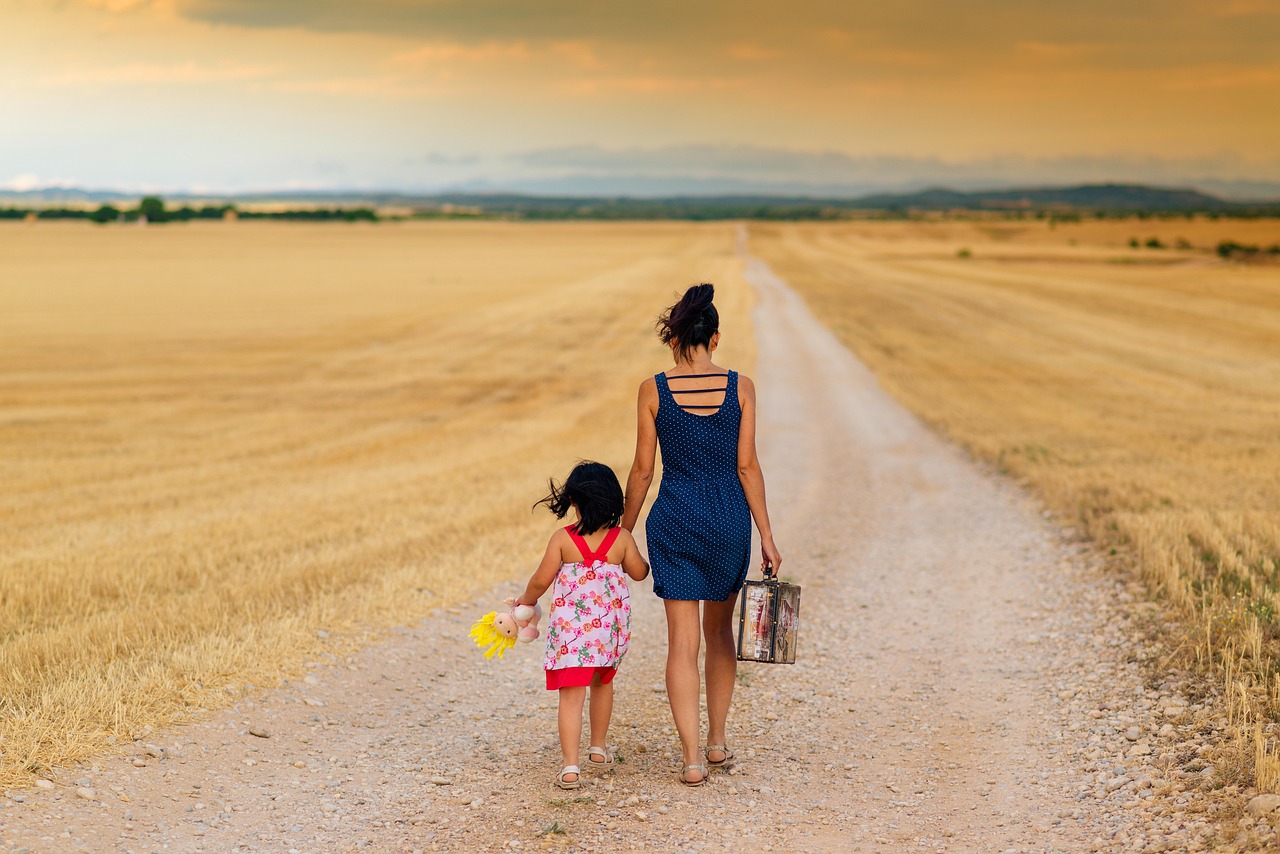 A woman and a child walk down a dirt path through a golden field, carrying a small suitcase.